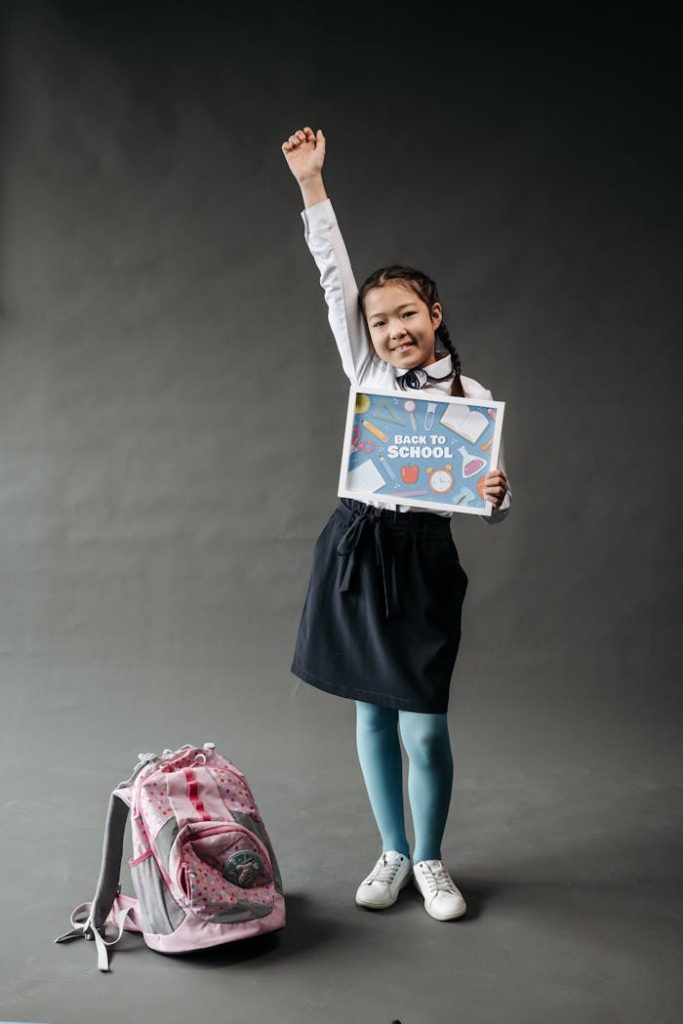 pexels-photo-8423894 Asian girl with backpack and 'Back to School' poster, raising her hand, studio shot.